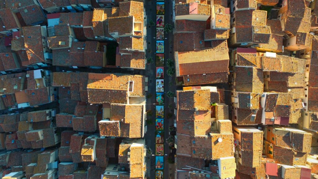 Aerial view of a traditional Sicilian village street with colorful artwork and terracotta-roofed houses on either side.